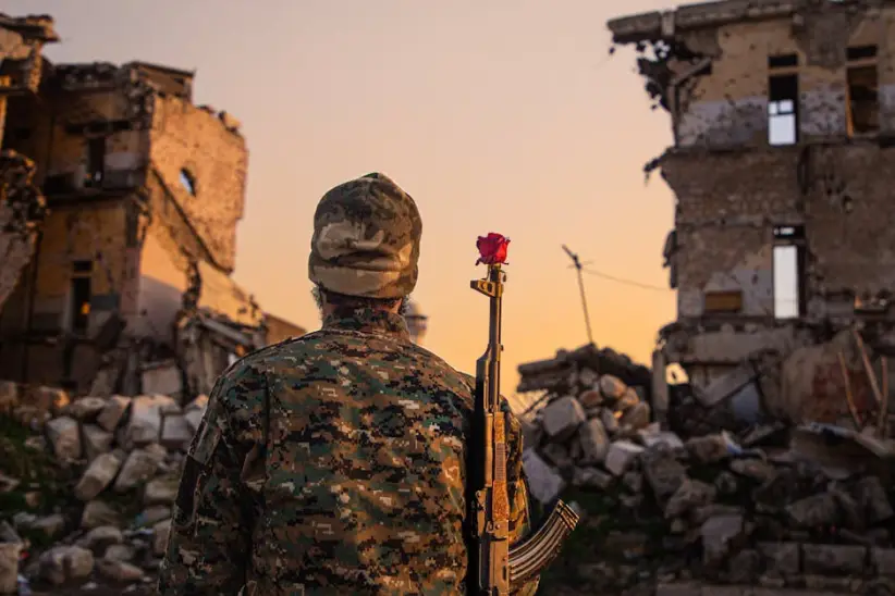 Un soldado camuflado con una rosa roja en un rifle en medio de edificios en ruinas al atardecer.