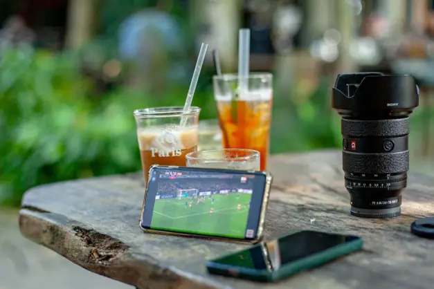 Un teléfono inteligente transmite fútbol junto a bebidas y equipos fotográficos en un café al aire libre.