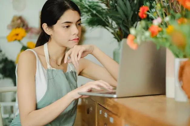 Una florista con un delantal trabaja en un portátil entre flores coloridas en una florería. (Foto: Pexels)