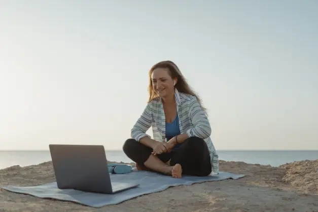 Mujer sentada en la playa usando un portátil mientras lleva auriculares inalámbricos, disfrutando de un entorno de trabajo relajante. (Foto: Pexels)