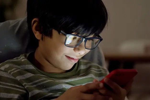 Niño joven usando un smartphone con gafas, sonriendo durante la noche en interiores. (Foto: Pexels)