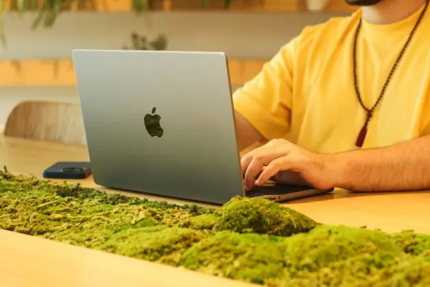 Hombre con camisa amarilla trabajando en laptop en una mesa de madera decorada con musgo en interiores. (Foto: Pexels)