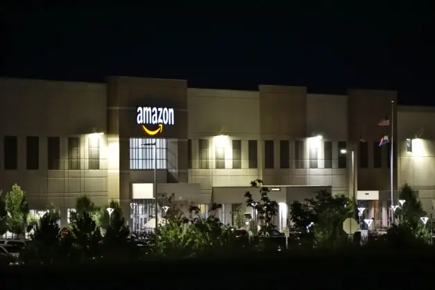 Edificio del almacén de Amazon iluminado por la noche con árboles y señalización. (Foto: Pexels)