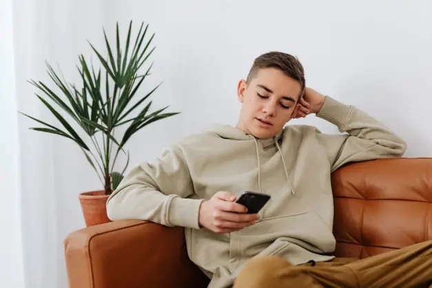 Chico adolescente sentado en casa enviando mensajes de texto en un smartphone, vistiendo una sudadera. (Foto: Pexels)