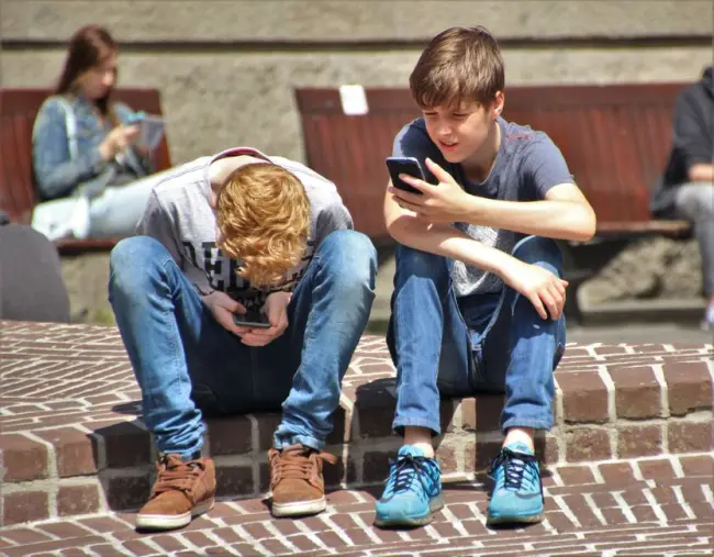 Dos adolescentes sentados al aire libre, enfocados en sus smartphones, abrazando la tecnología moderna. (Foto: Pexels)