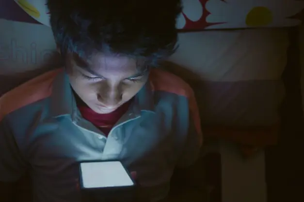 Niño acostado en la cama mirando atentamente la pantalla brillante del smartphone. (Foto: Pexels)