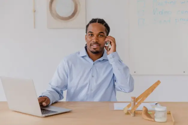 Joven en escritorio con smartphone, usando laptop, sonriendo. Entorno de trabajo profesional. (Foto: Pexels)