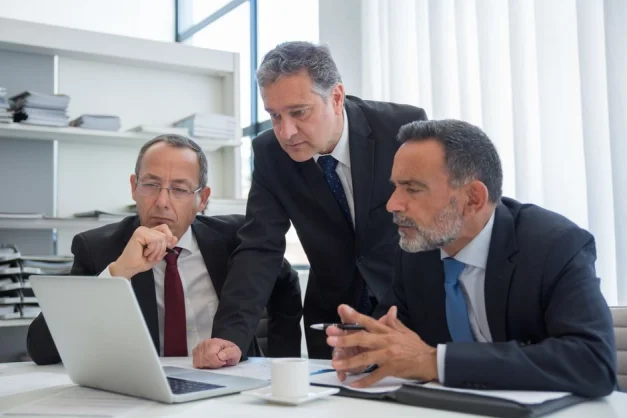 Tres empresarios en trajes colaborando en un entorno de oficina moderno, centrados en un portátil.