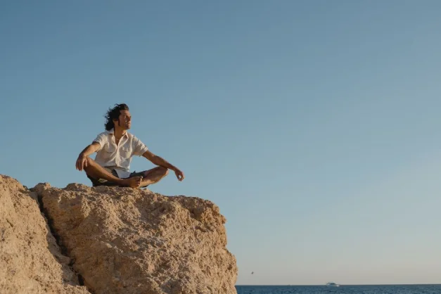 Hombre en posición de meditación en un acantilado rocoso con vista al océano al atardecer, abrazando la serenidad y la naturaleza.
