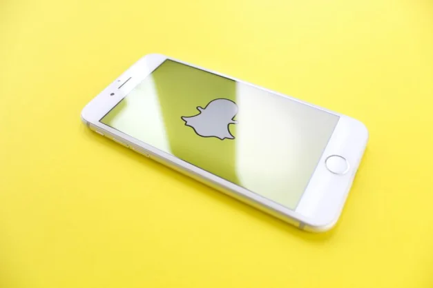 Smartphone blanco mostrando el logo de Snapchat sobre un fondo amarillo brillante, vista en ángulo.