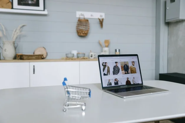 Una laptop que muestra un portal de compras en línea se encuentra en un escritorio minimalista con un carrito de compras en miniatura en primer plano.