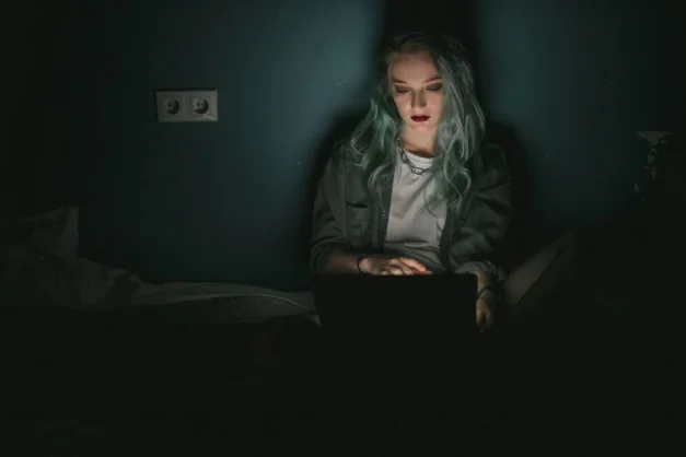 Una mujer con el cabello teñido usando un portátil en una habitación poco iluminada, trabajando tarde en la noche.