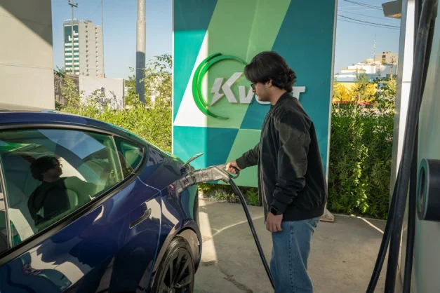 Joven cargando un Tesla Model 3 en una estación de vehículos eléctricos al aire libre durante el día.
